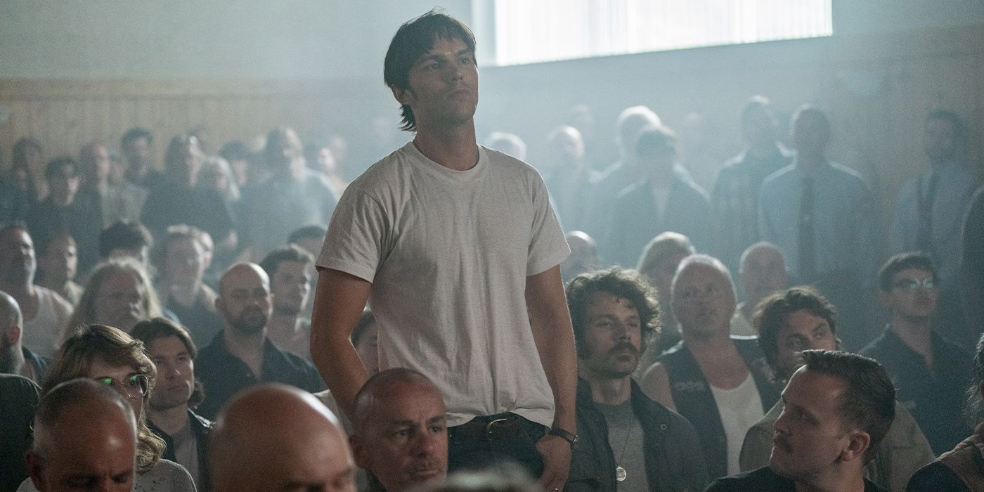 Mathews (Nicholas Hoult) standing in a crowd of seated people with his hands in his pockets