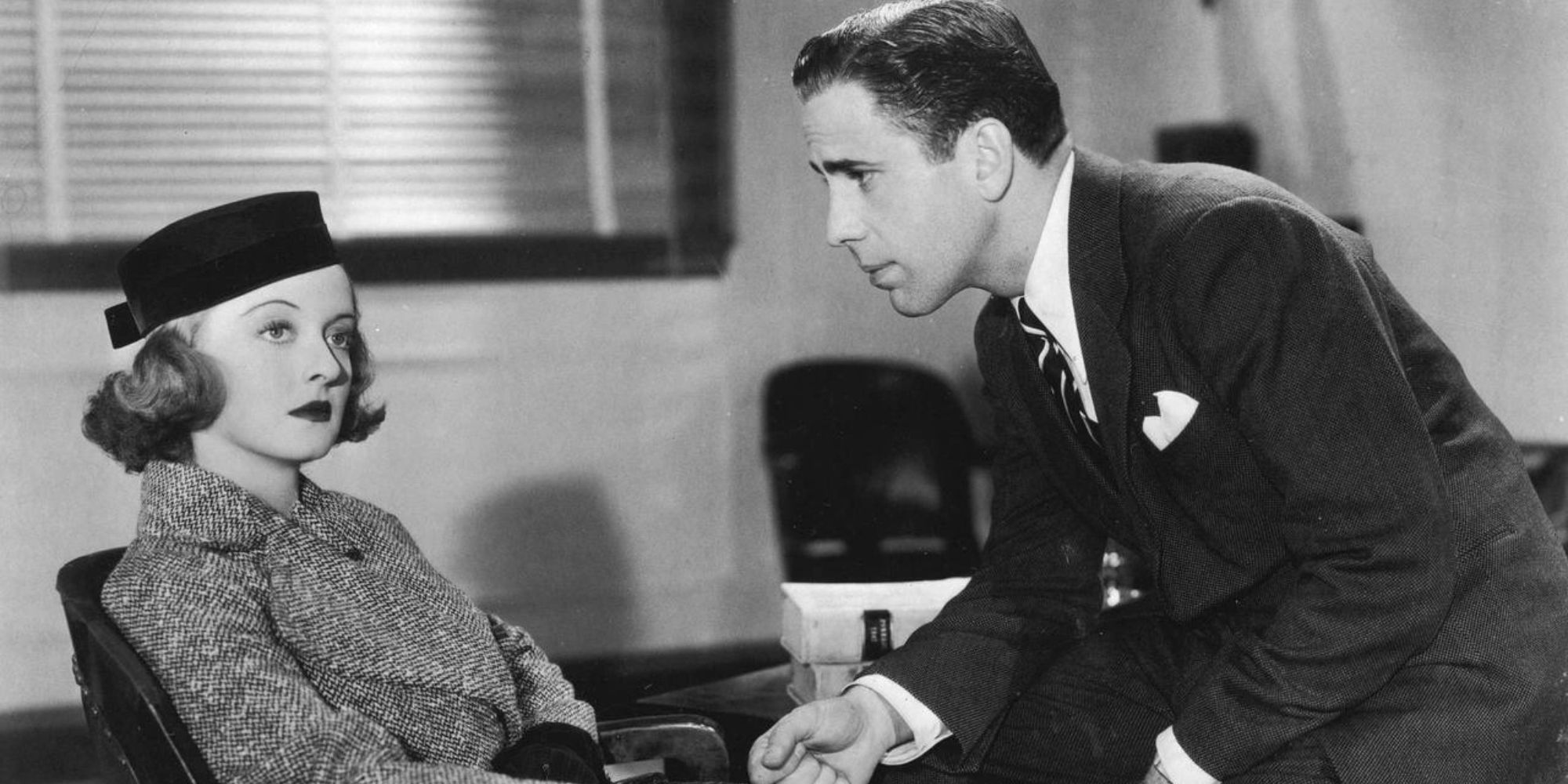 Bette Davis looking to the side while sitting in a chair across from Humphrey Bogart who is staring at her in Marked Woman (1937)