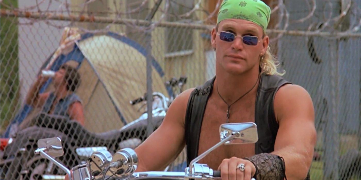 A man in a vest, green bandana, and blue sunglasses is sitting on a motorcycle and holding the wheel while looking over his glasses at something off-screen