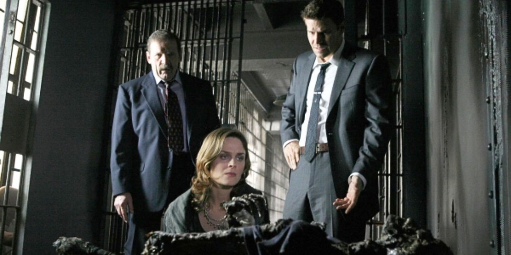 David Boreanaz and Mik Scriba standing behind Emily Deschanel as she is kneeling next to a decomposed body in a prison cell in Bones episode The Man in the Cell
