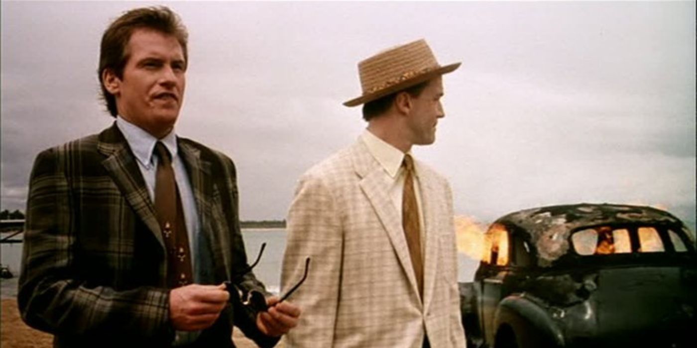 Denis Leary and Douglas McGrath stand on a beach in suits while a car burns behind them in Company Man