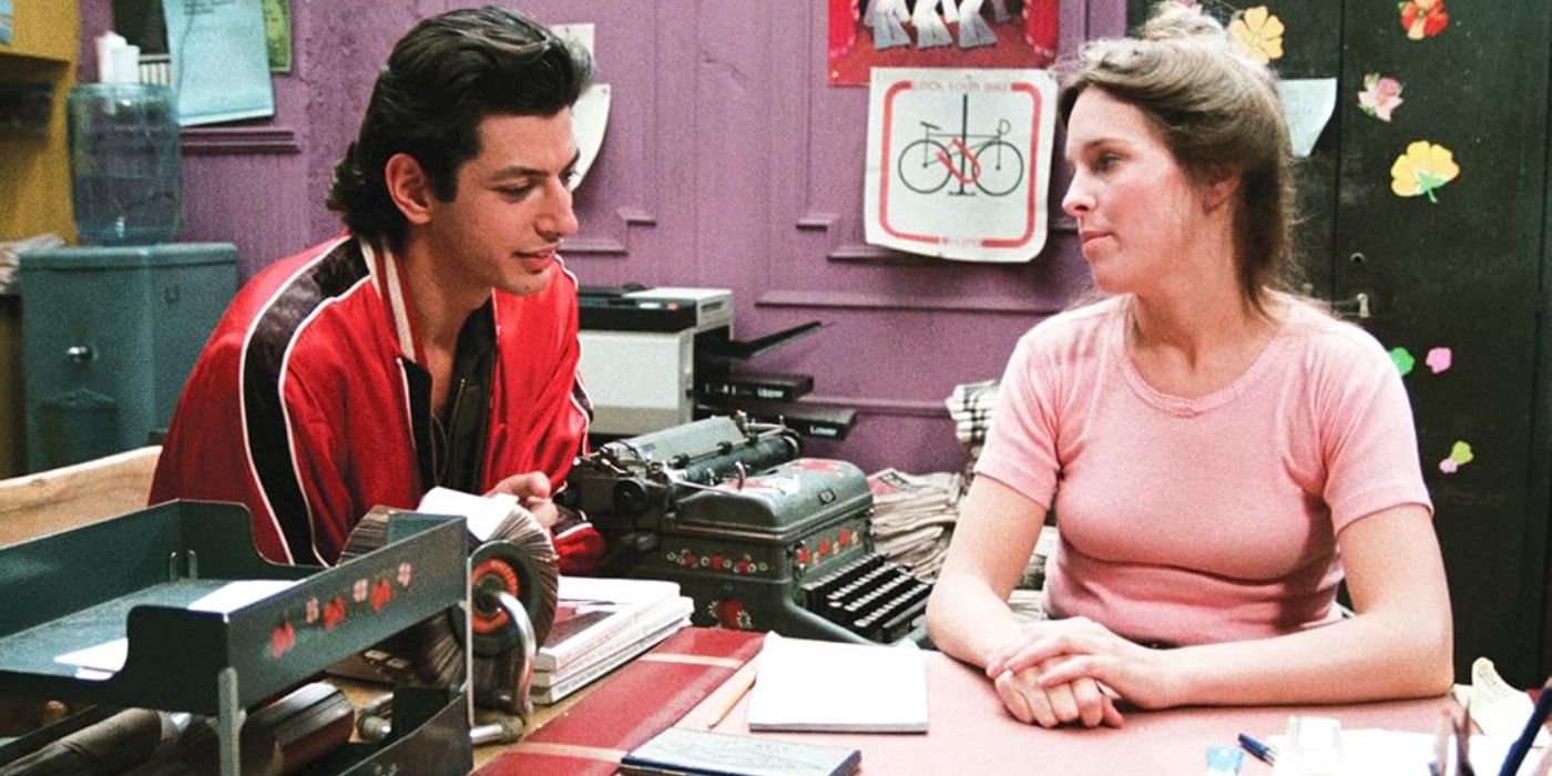 Jeff Goldblum in a red and black jacket sits at a desk talking to a woman in a pink shirt in Between the Lines.