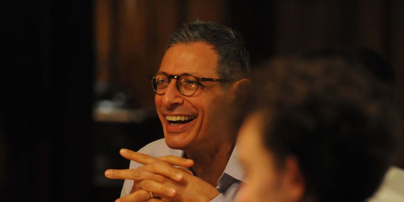 Jeff Goldblum with fingers interlaced laughing and looking at someone off-screen in Le Week-End.