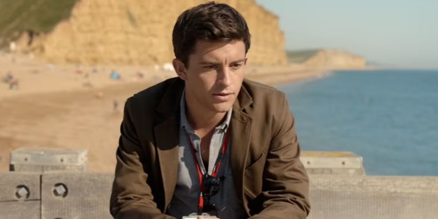 Jonathan Bailey with a badge around his neck, sitting and looking at something on a beach in Broadchurch.