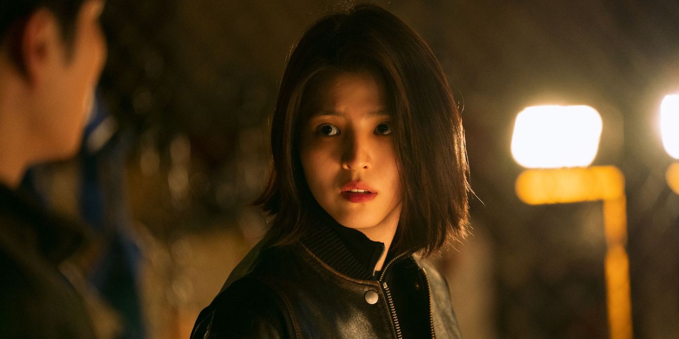 Ji-woo (Han So-hee), wearing a black leather jacket and with short bobbed hair, looking toward the left at a man who's mosty offscreen in My Name