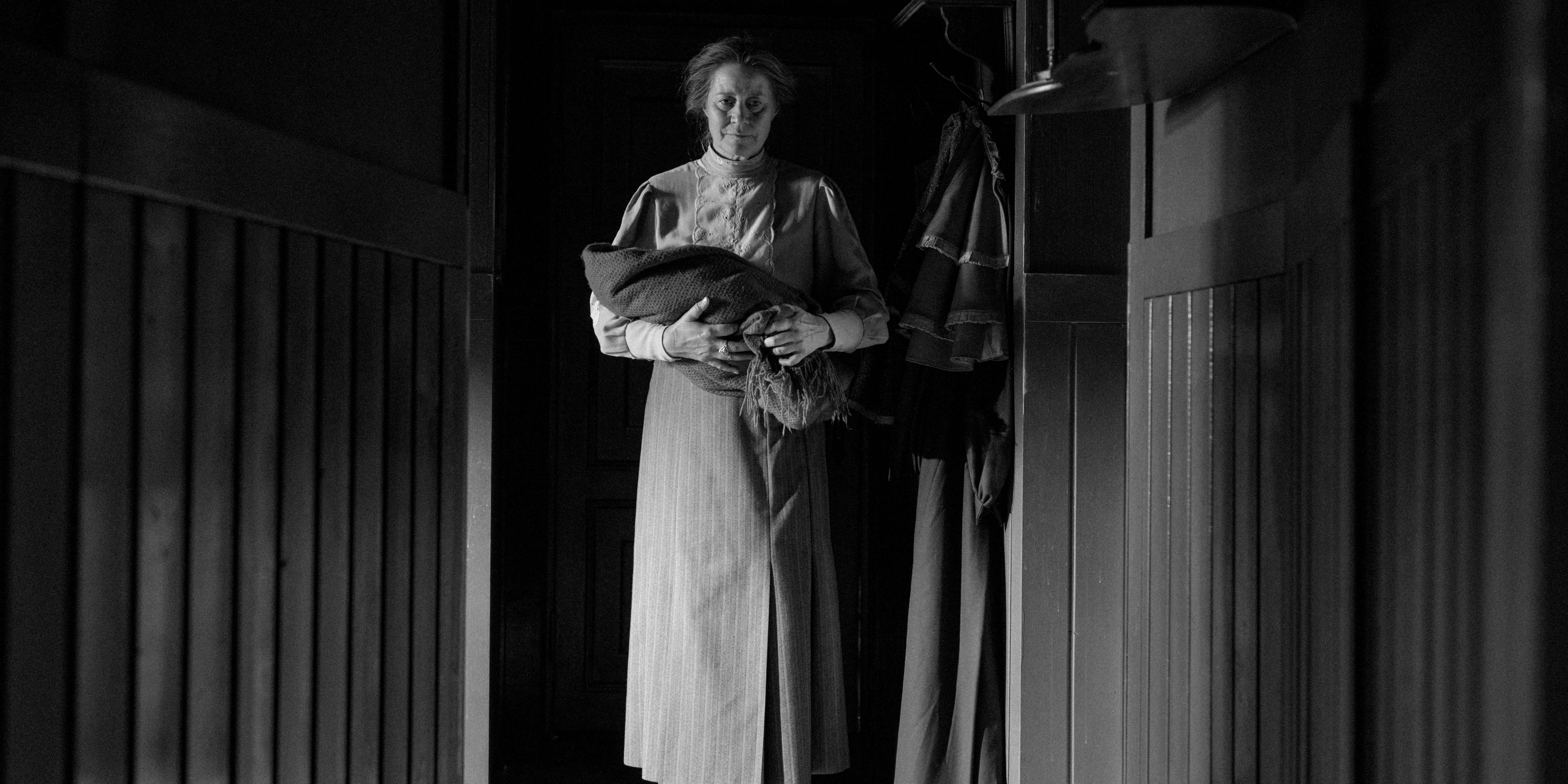 Dagmar, played by actor Trine Dyrholm, stands in a dark hallway holding a baby in The Girl With The Needle.