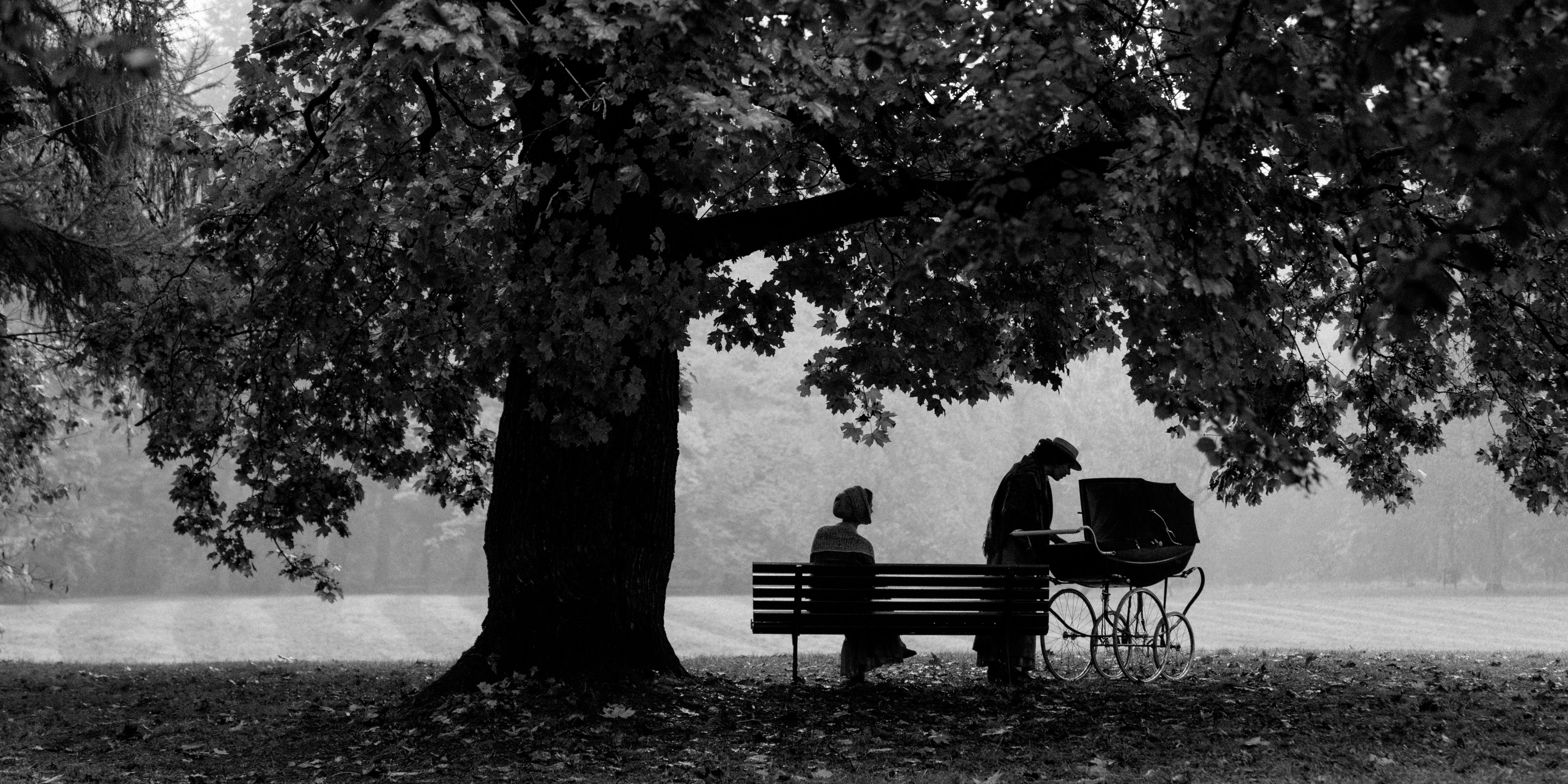 The figures of two women under a tree next to a baby carriage in the Girl With The Needle