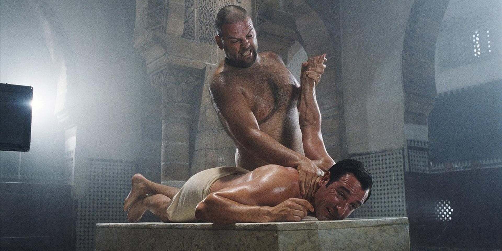 A man recoiling in main as he receives a painful massage from a shirtless man in 'OSS 117: Cairo, Nest of Spies'