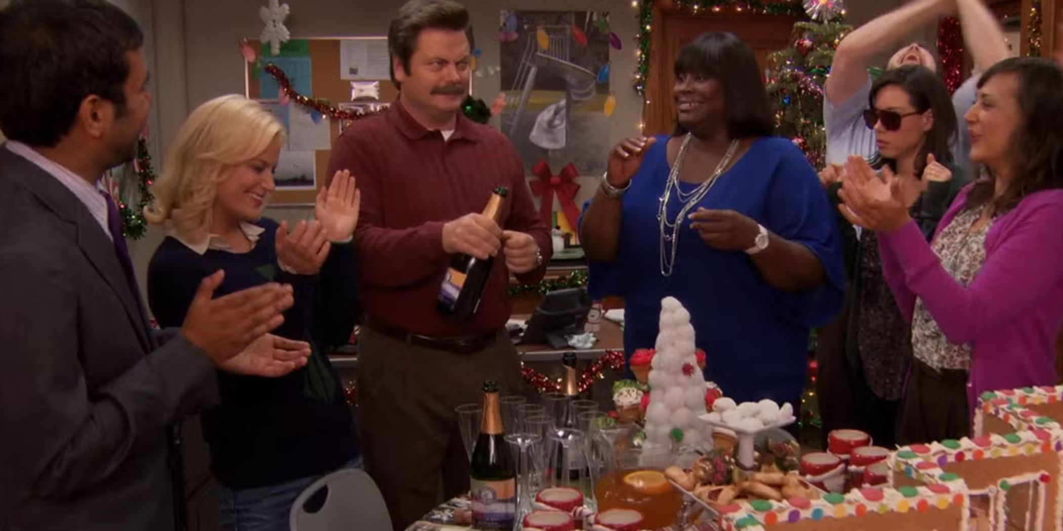 Tom (Aziz Ansari), Leslie (Amy Poehler), Ron (Nick Offerman), Donna (Retta) Andy (Chris Pratt), April, (Aubrey Plaza), and Ann (Rashida Jones) celebrating around a gingerbread house in Parks and Recreation