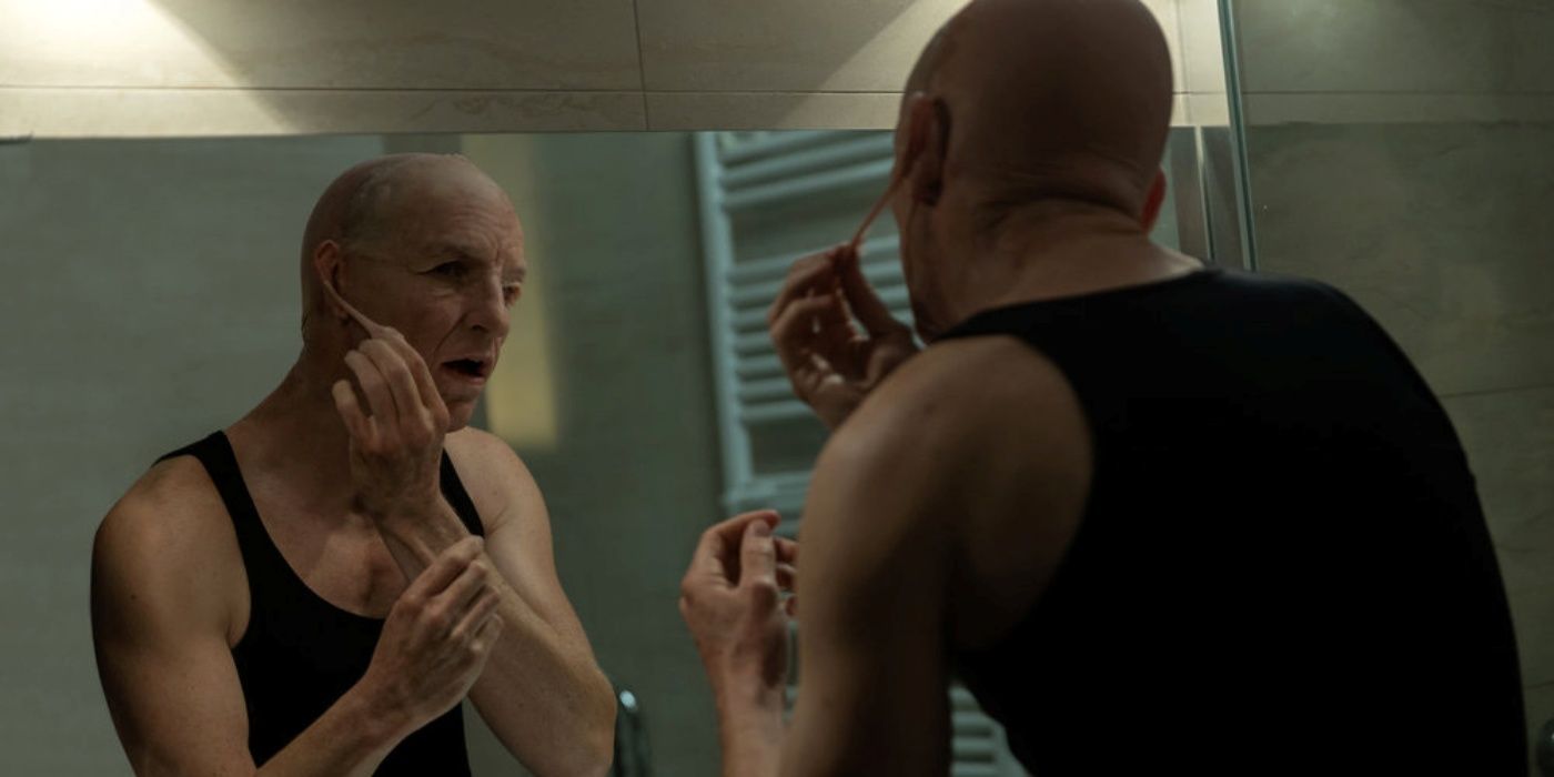 Eddie Redmayne as The Jackal peeling off the face of one of his disguises in the mirror in The Day of the Jackal