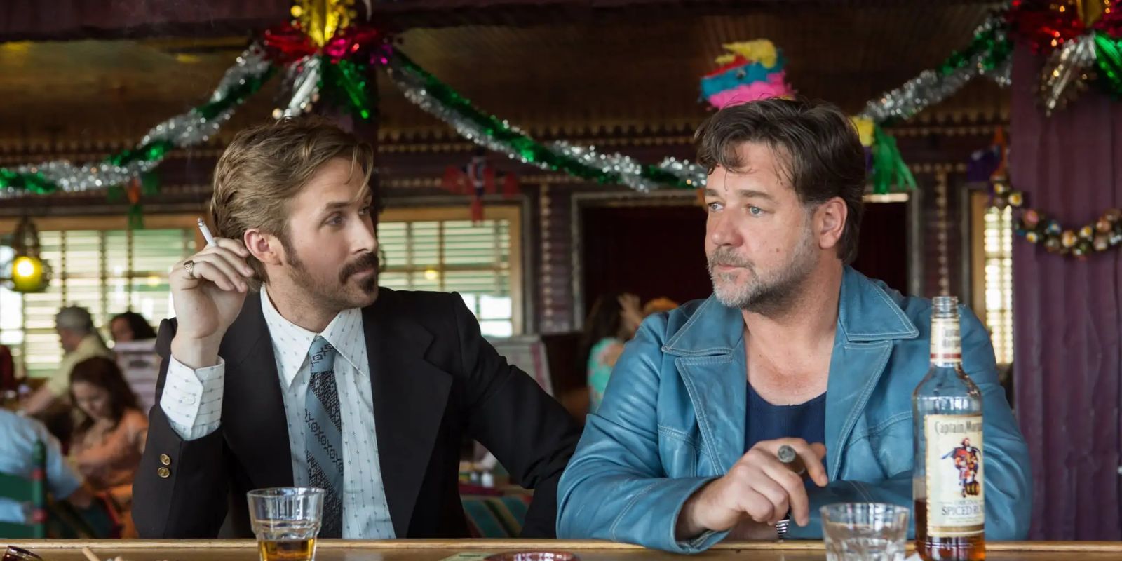 Two men, Holland March and Jackson Healy, smoke and drink while seated at a bar with Christmas decorations