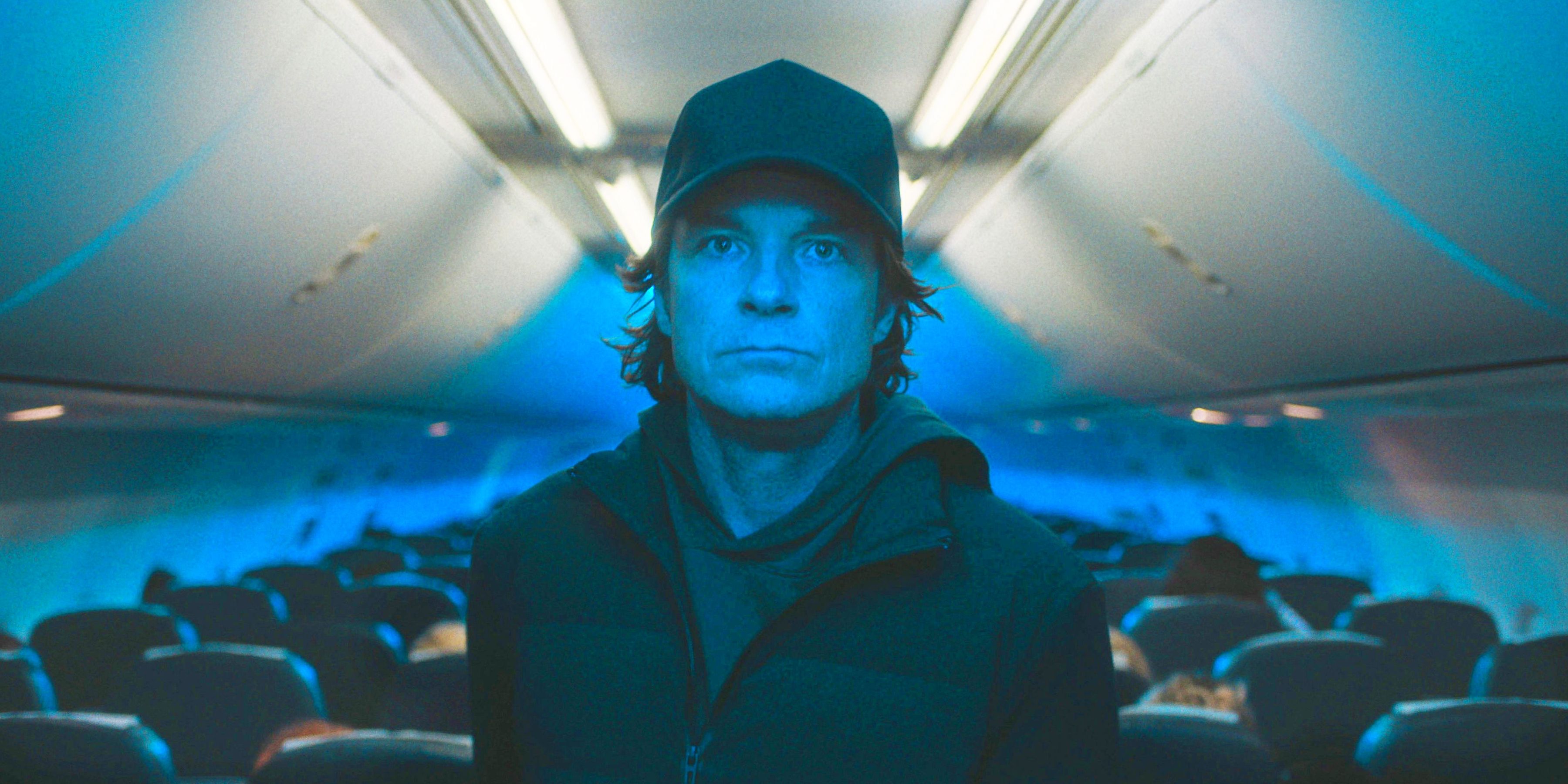 Jason Bateman as The Traveler, standing ominously in the aisle of an airplane wearing a black baseball cap and bathed in blue light.