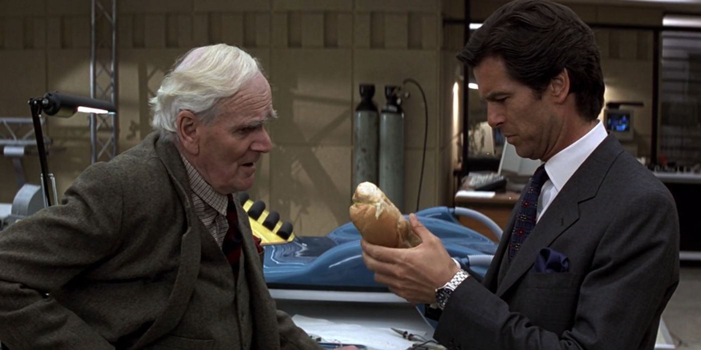 Desmond Llewelyn as Q and Pierce Brosnan as Bond talking about weapons while Bond is holding a large sandwich in his left hand