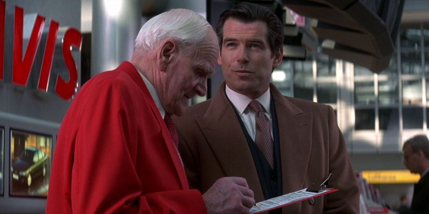 Desmond Llewelyn as Q and Pierce Brosnan as James Bond in 'Tomorrow Never Dies', with Q writing something on a piece of paper attached to a clipboard and Bond looking off-screen