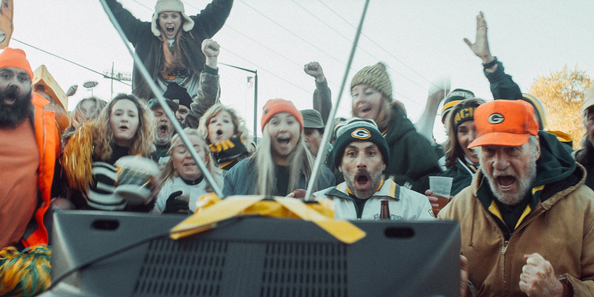 A group of Packers fans cheer around a television set while wearing the team colors in Green and Gold.