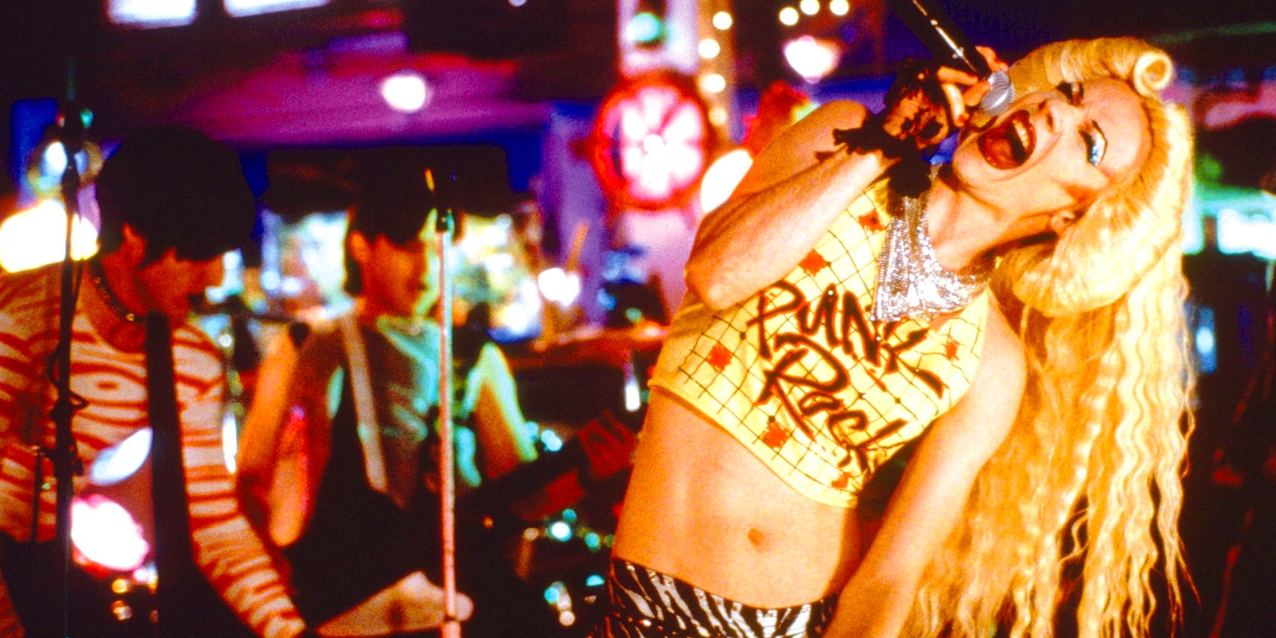 Hedwig, played by actor John Cameron Mitchell, howling into a microphone on stage while guitarists play behind her.