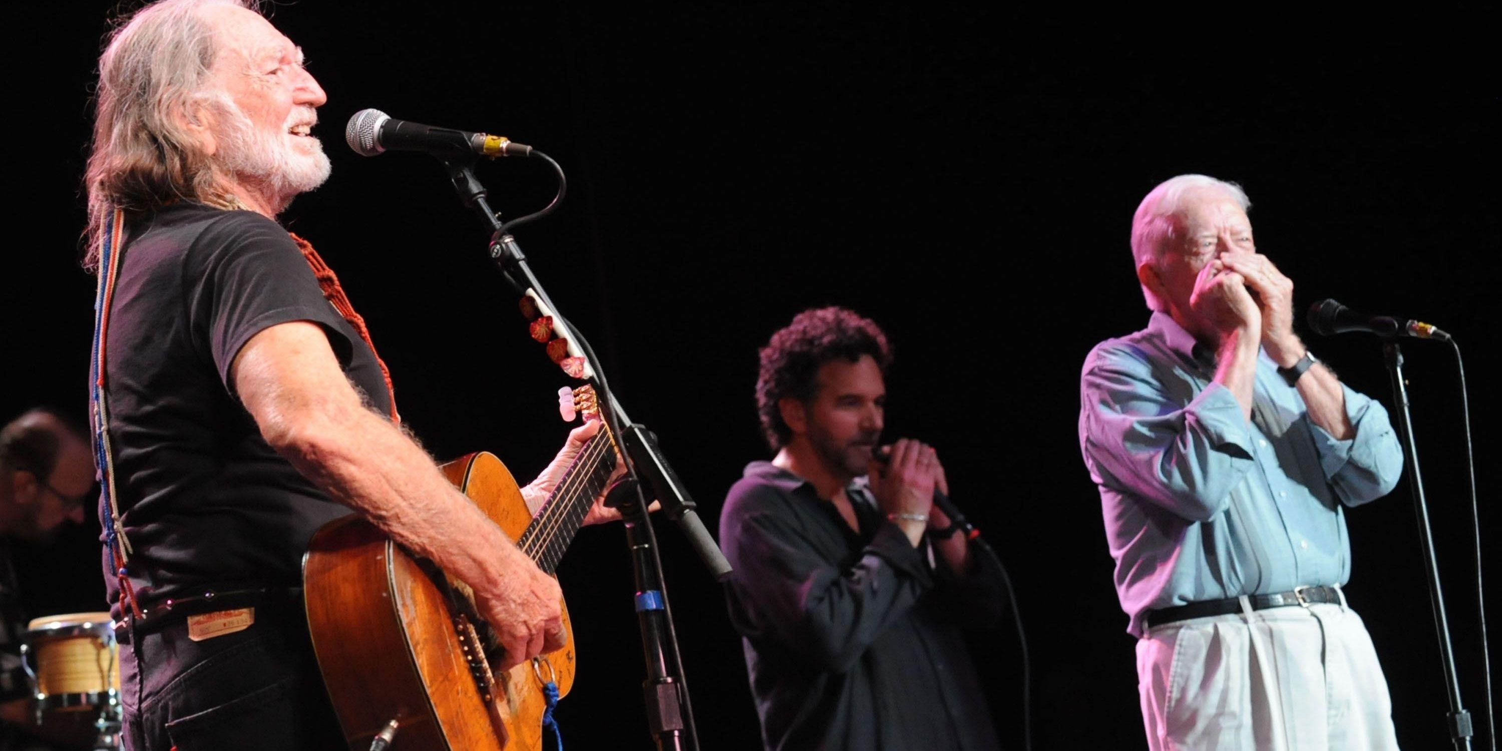 Jimmy Carter playing a harmonica on stage next to Willie Nelson, who is playing a guitar.