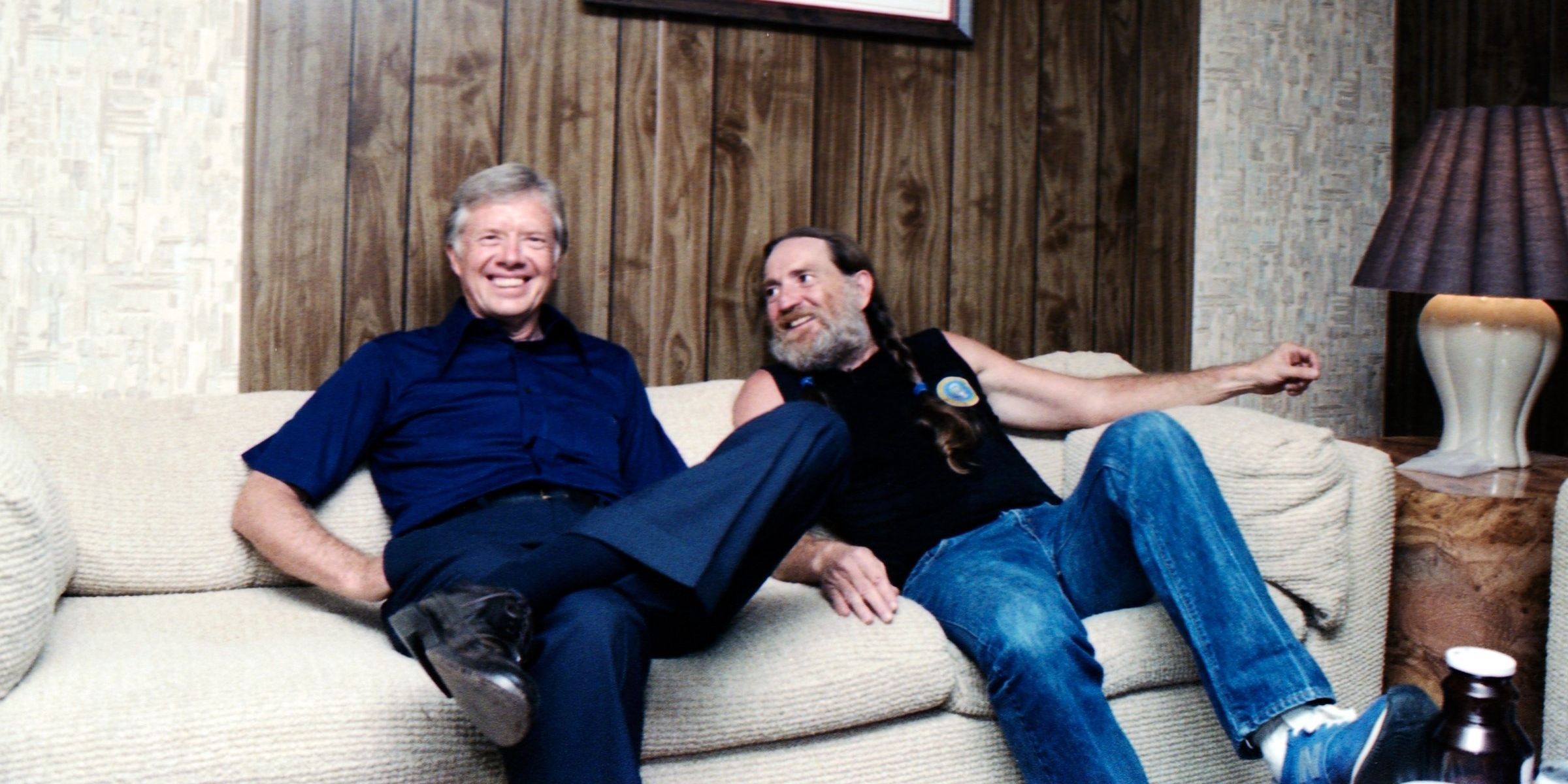 Jimmy Carter sat smiling on a couch next to musician Willie Nelson.