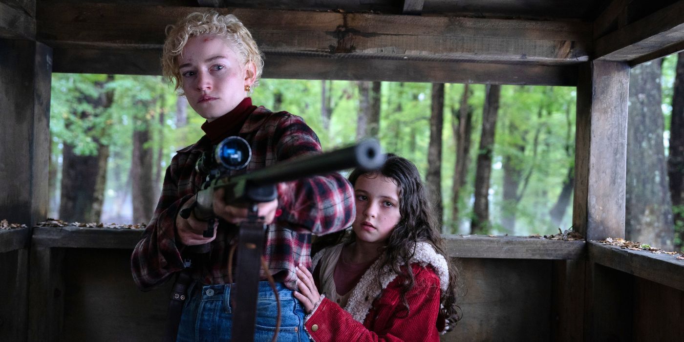 Julia Garner as Charlotte, holding a sniper rifle, while Matilda Firth as Ginger, stands behind her, in Wolf Man.