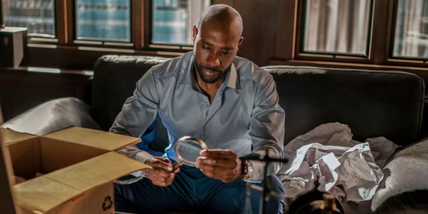 Morris Chestnut plays Dr John Watson in CBS show. He is seated on a couch in a dimly lit room examines a magnifying glass with focused attention. He is wearing a light blue shirt, with an open cardboard box and crumpled paper nearby, suggesting an unpacking or investigative scene. Large windows in the background provide a view of a cityscape.