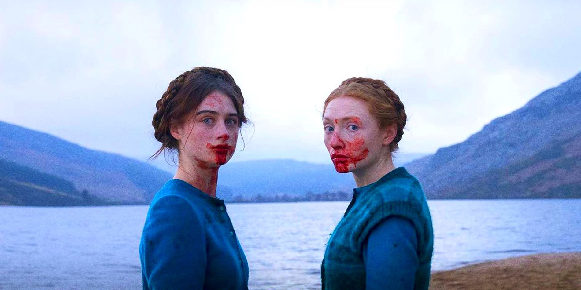 Two young women stand next to open water wearing identical blue dresses, with blood smeared on their faces.