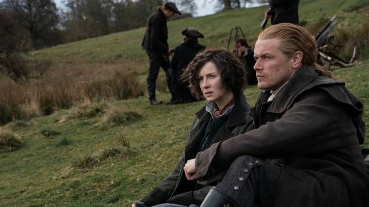 Caitriona Balfe as Claire and Sam Heughan as Jamie sit on a hillside in period clothes in Outlander.