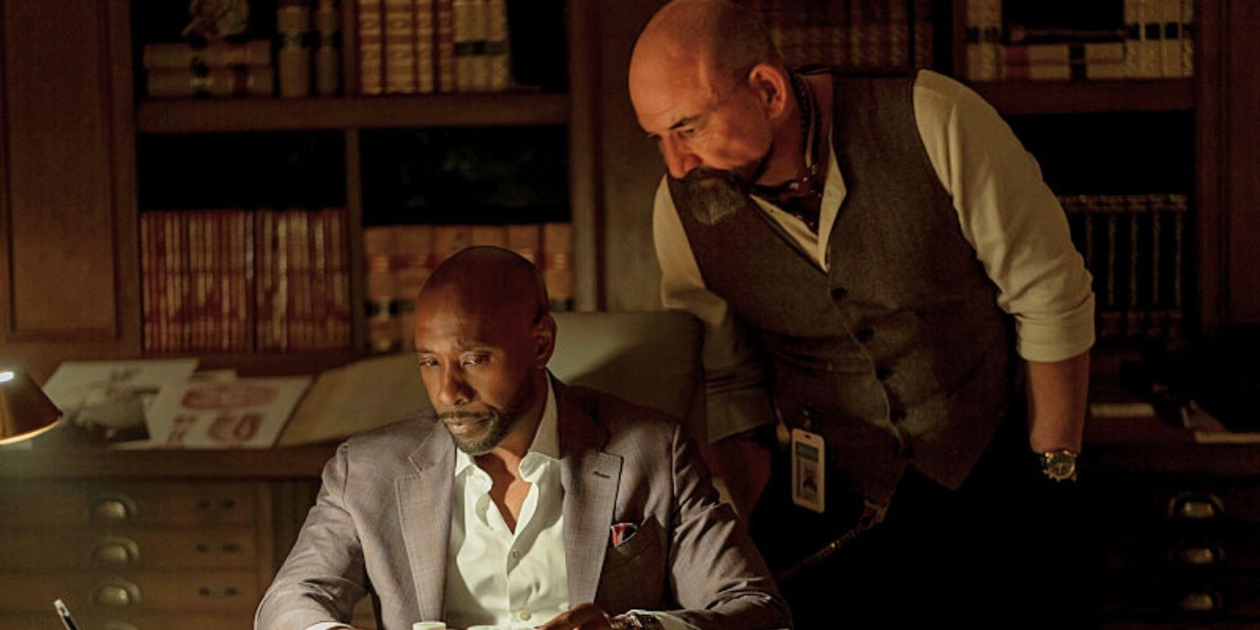 Ritchie Coster as Shinwell Johnson and Morris Chestnut as Dr. John Watson in the CBS show Watson. Morris Chestnut sits at a desk, intently focused on documents under a desk lamp, while Ritchie Coster stands beside him.