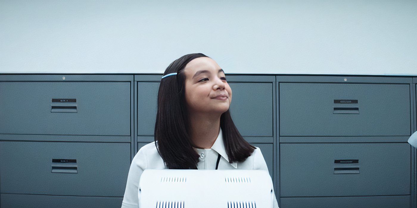 Miss Huang (Sarah Bock) politely smiles at some passing employees from her desk in Apple TV&rsquo;s &lsquo;Severance&rsquo;.