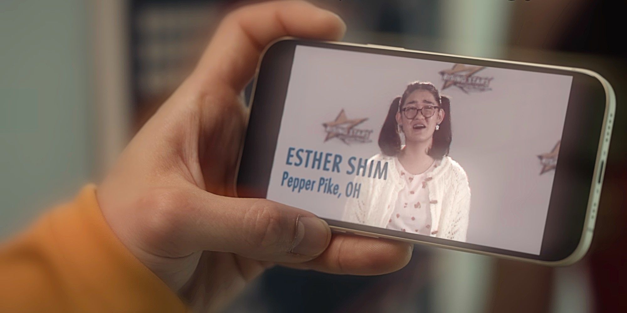 Minho's hand holding a cell phone in XO, Kitty Season 2. On the screen in Stella from many years before. Underneath her, it reads: "ESTHER SHIM Pepper Pike, OH."