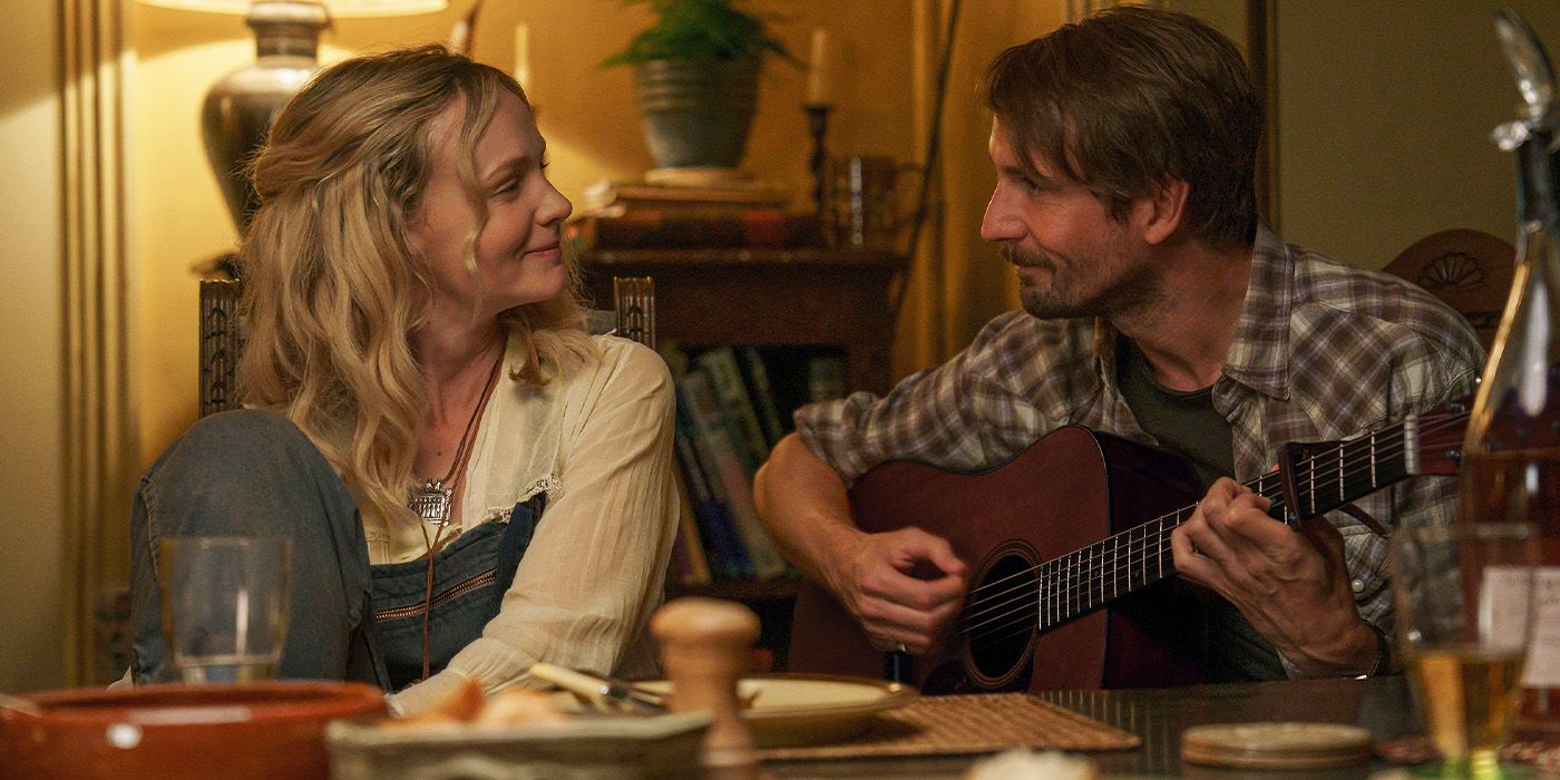 Carey Mulligan smiling at Tom Basden playing guitar at a table in The Ballad of Wallis Island