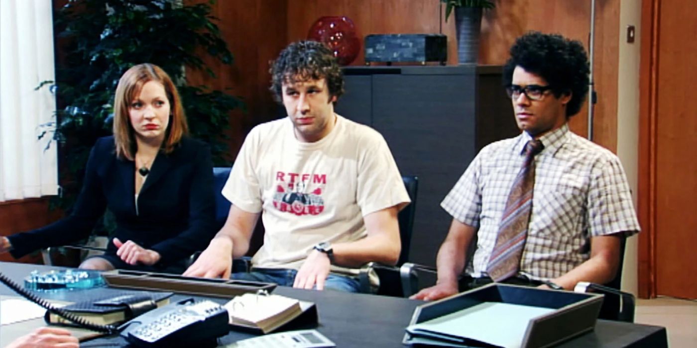 The cast of The IT Crowd sit awkwardly in front of a desk in an office building. 