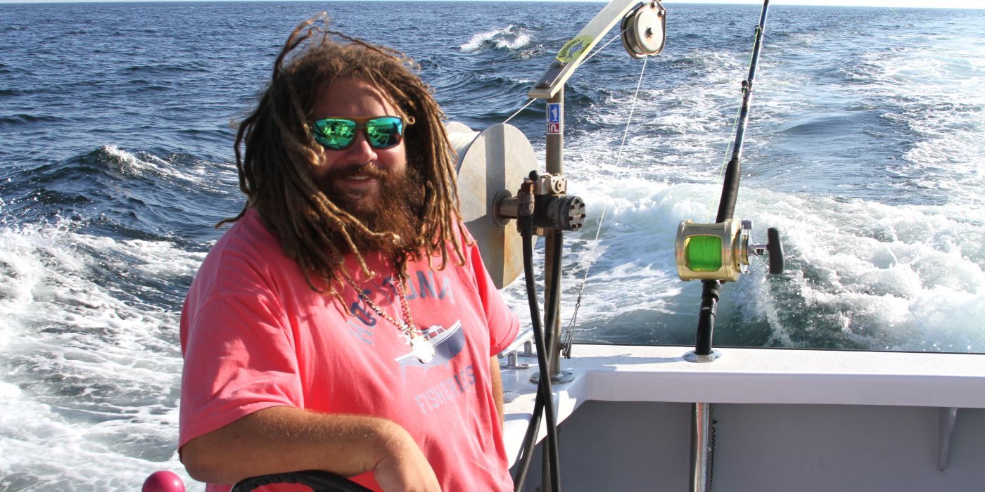 JT Ott in sunglasses smiling as he stands on his boat as it sails on water in an episode of Wicked Tuna.