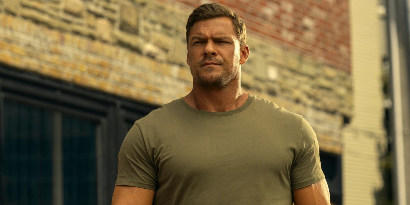 Alan Ritchson as Jack Reacher, wearing a beige t-shirt, standing in front of a brick building.