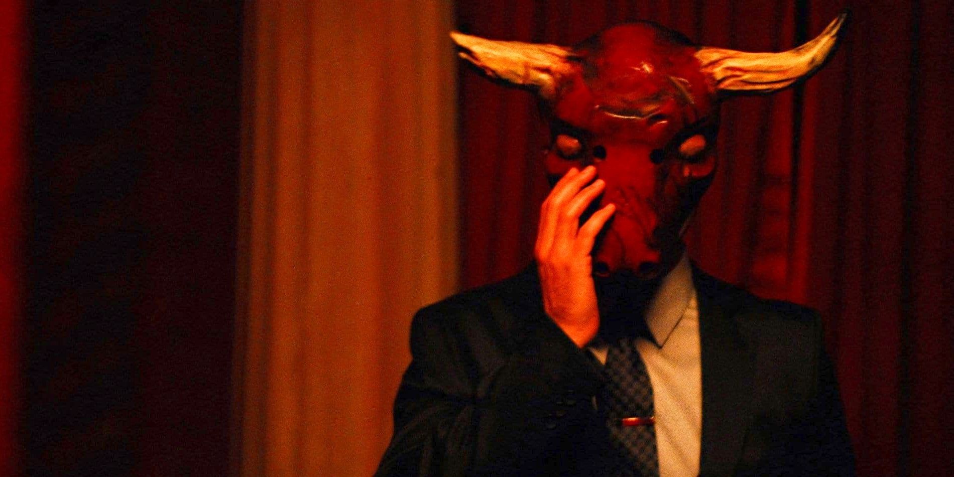 A man in a suit wearing a menacing bull mask puts his hand to his face in The Conspiracy.