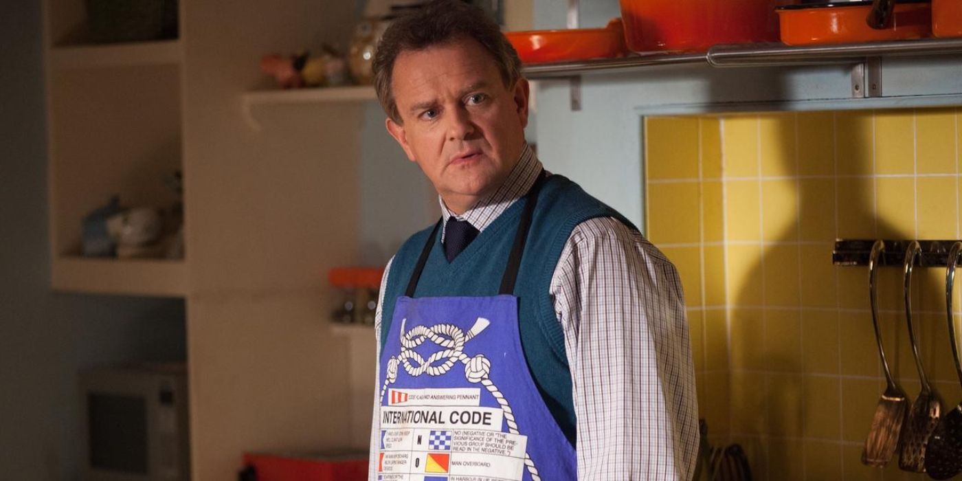 Hugh Bonneville as Mr. Brown wears an apron and stands in the kitchen in the Paddington film series.