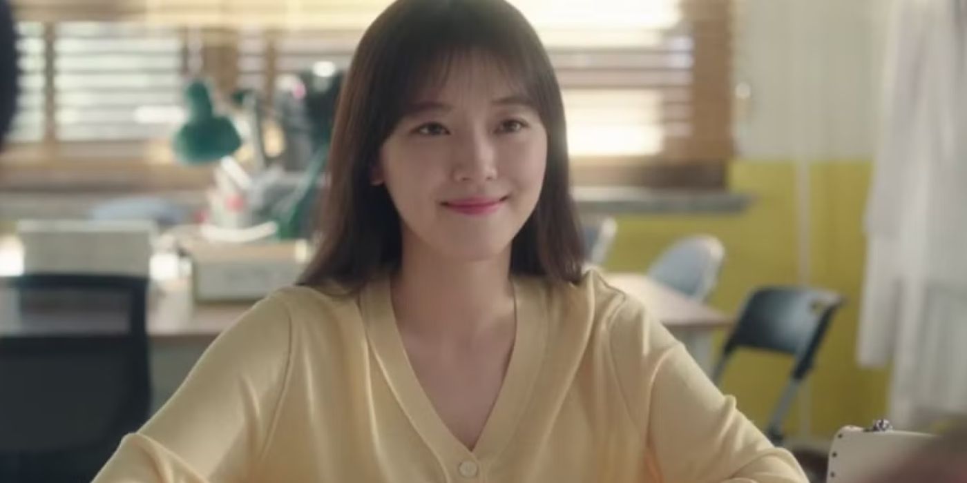 Jo Woo-ri as Hyun Soo-ah in a yellow sweater, at a desk and smiling at people in front of her in Gangnam Beauty.