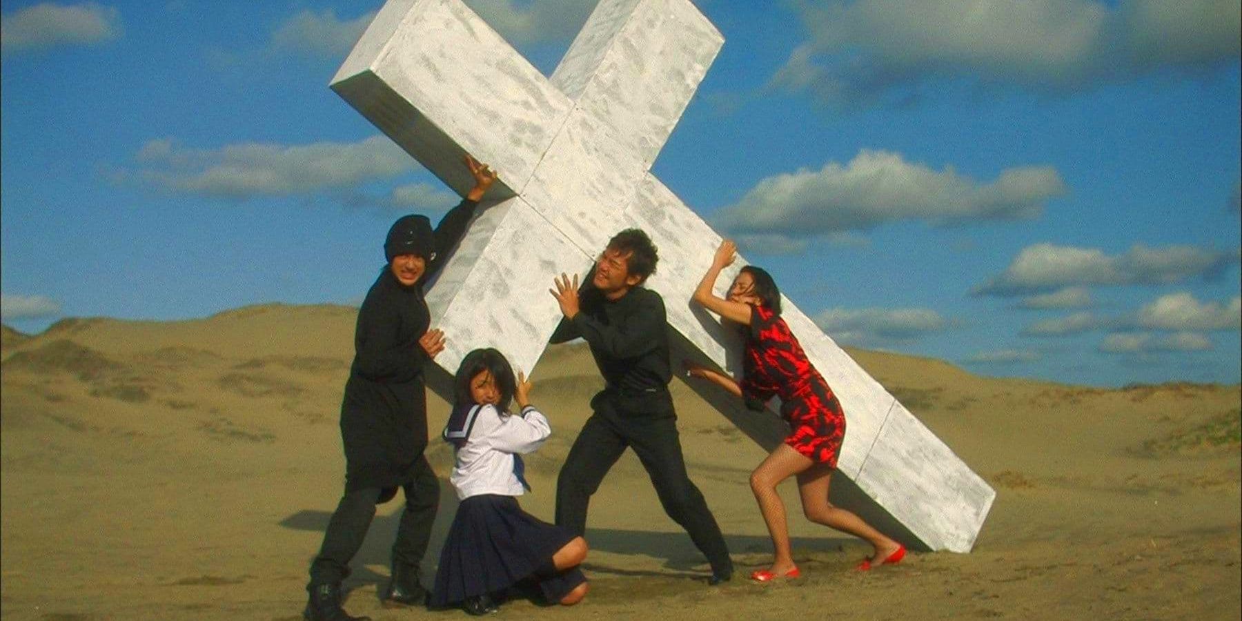 Two men and two women struggle to hold up a giant white crucifix in the desert in Love Exposure.