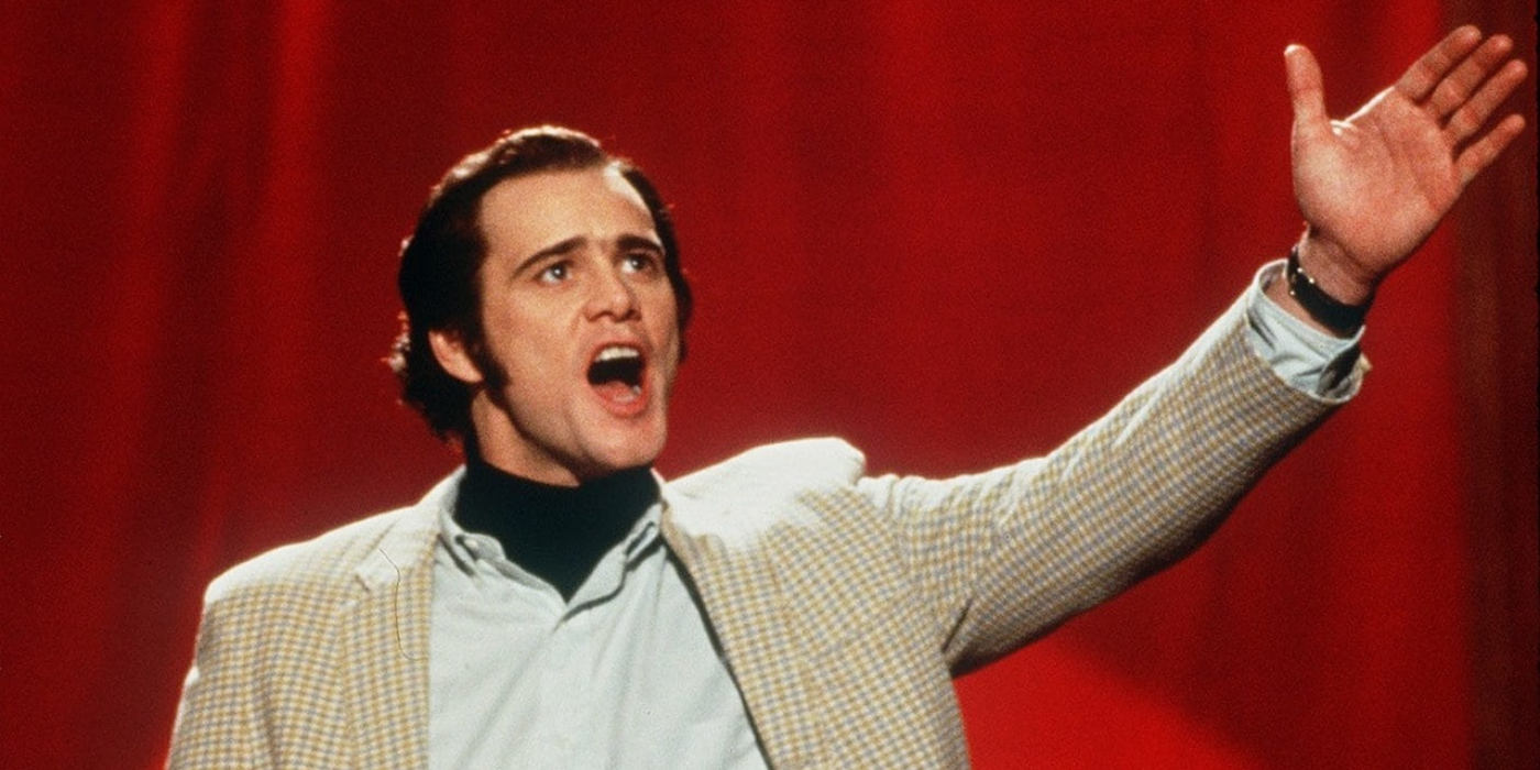 Jim Carrey stands against a red curtain with his hand up