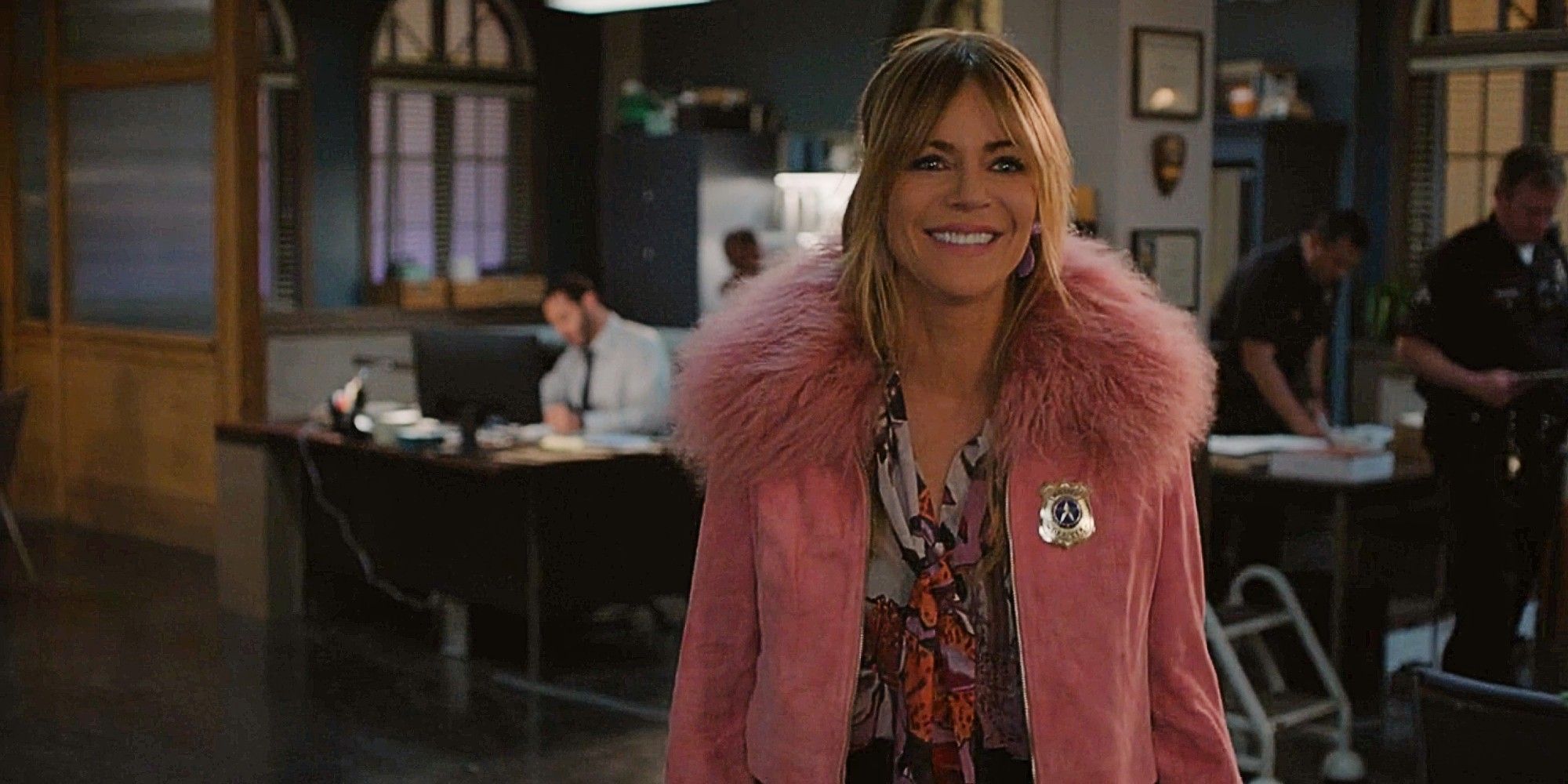 Morgan Gillory smiling and wearing a police badge on her jacket while Karadec works at a desk behind her in High Potential Season 1 Episode 11