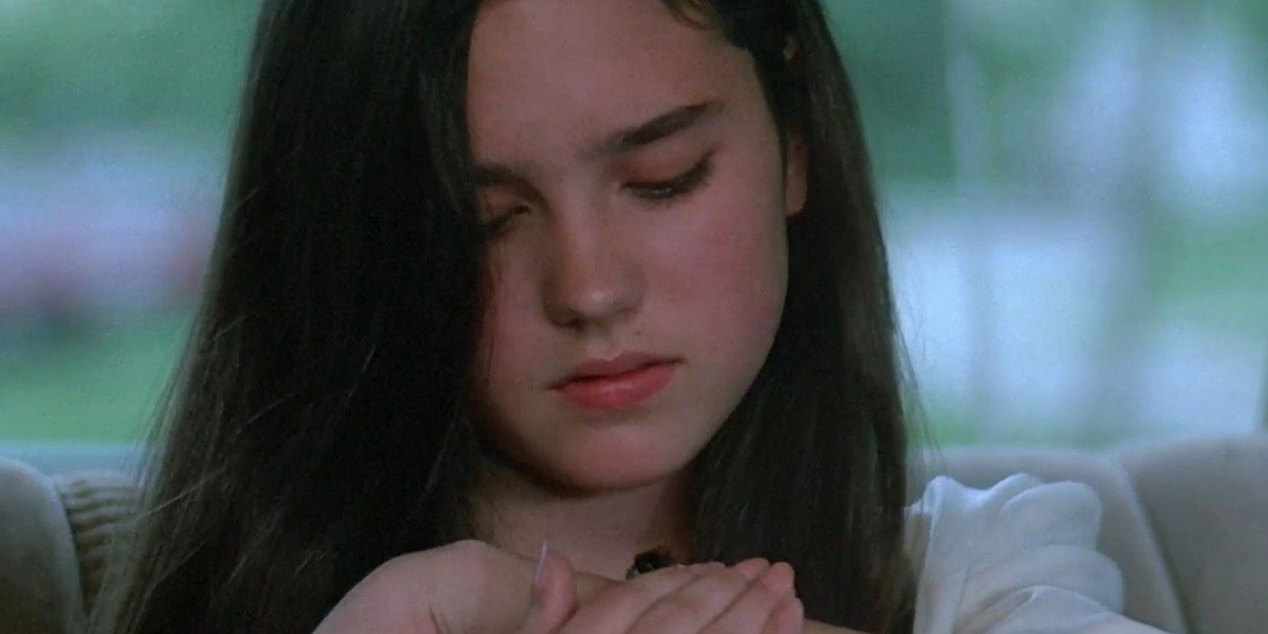 Jennifer Connelly wearing a white shirt, sitting in a car while protecting a bee on her hands in Phenomena.
