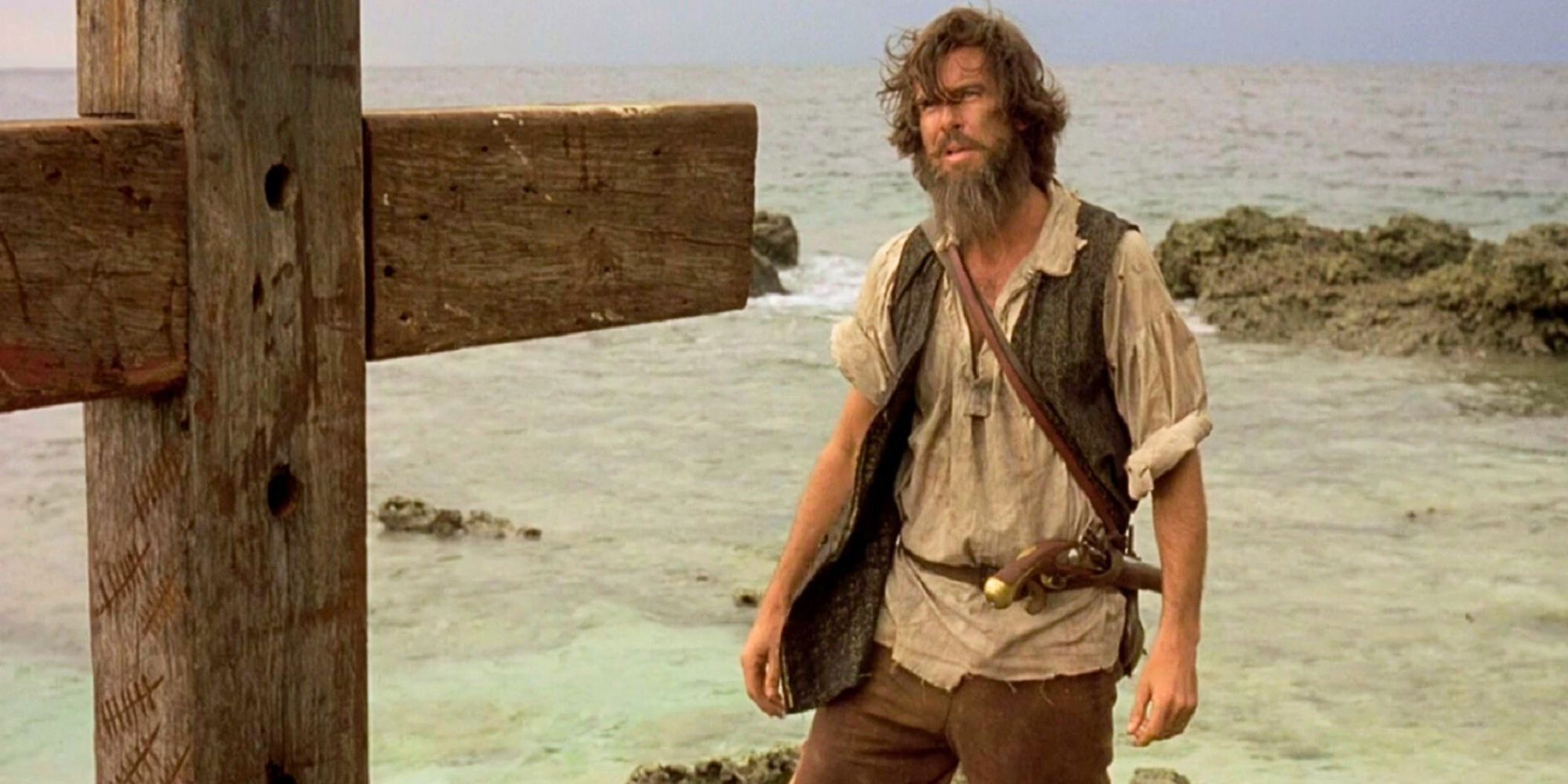 Pierce Brosnan standing on a beach looking at a large wooden cross in Robinson Crusoe (1997)
