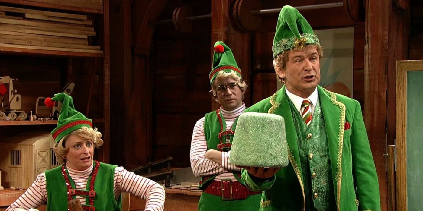 Alec Baldwin holding up a green ornament with two people dressed as elves behind him on SNL.