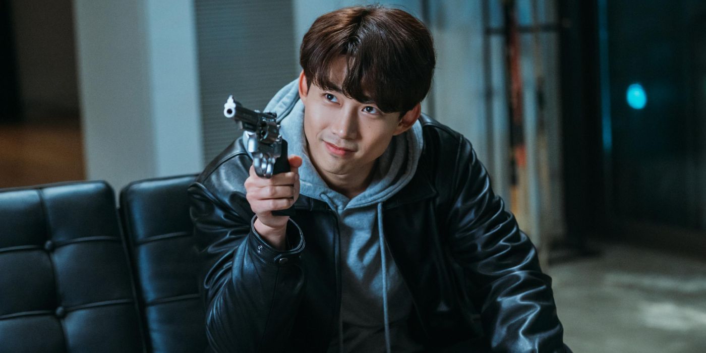 Ok Taecyeon in a leather jacket and gray hoodie smiles and points a gun at someone off-screen in Vincenzo.