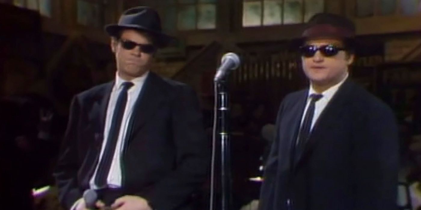 Dan Aykroyd and John Belushi on stage in suits with hats and sunglasses and a mic in front of them on SNL as The Blues Brothers.