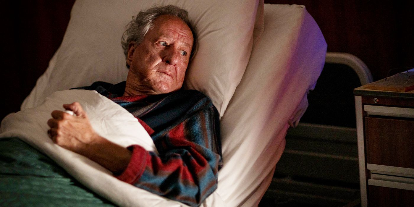 Geoffrey Rush in a red and blue robe under the covers of a hospital bed looking off to the side in fear