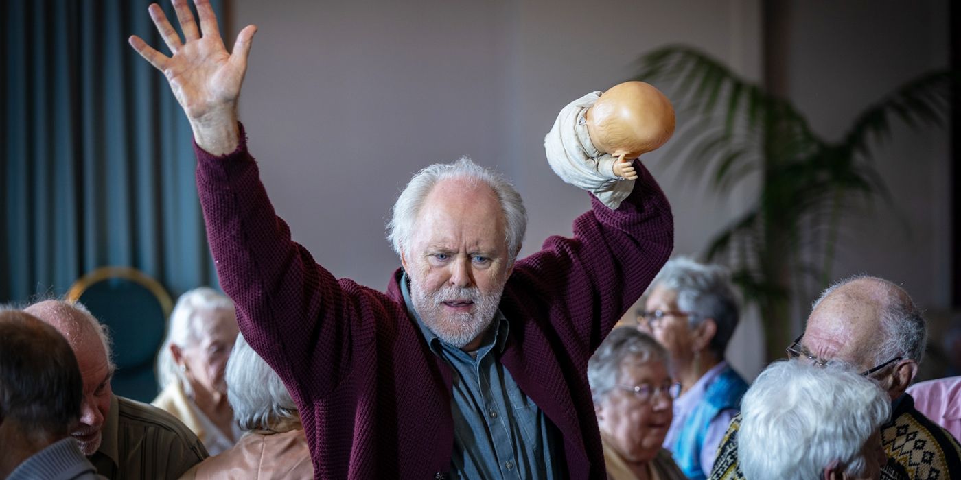 John Lithgow in a maroon swater holding up a toy doll in a crowd of elderly people looking focused