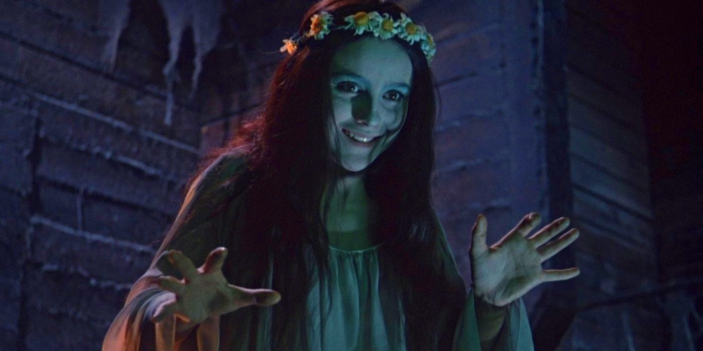 Natalya Varley as Pannochka, wearing a white robe and flower crown, holding her hands out spookily in Viy.