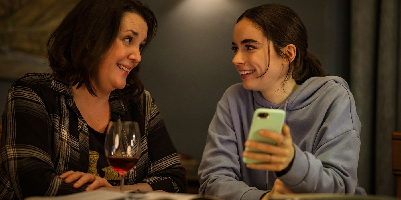 Melanie Lynskey and Sarah Desjardins as Shauna and Callie, smiling as Callie shows Shauna something on her phone in Yellowjackets Season 3