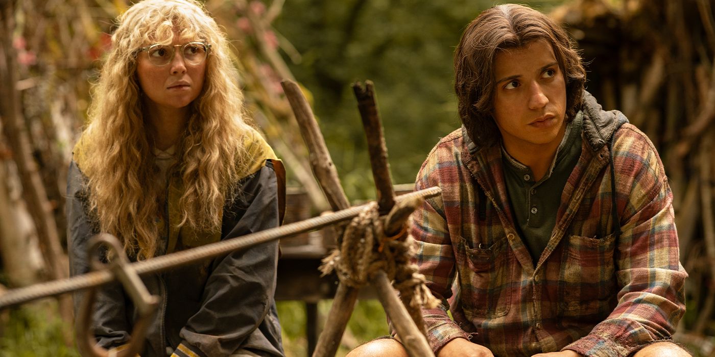 Misty and Travis sitting together at a fire pit looking to the side anxiously in Yellowjackets Season 3