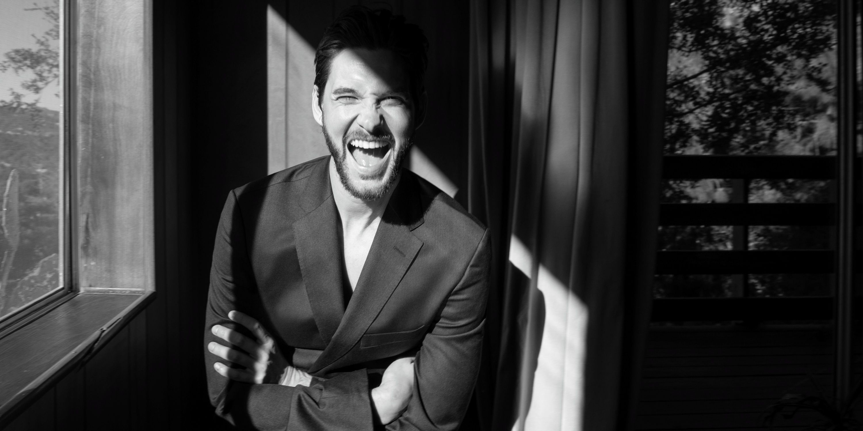 Ben Barnes in black and white laughing with his arms cross while leaning on a wall next to a window